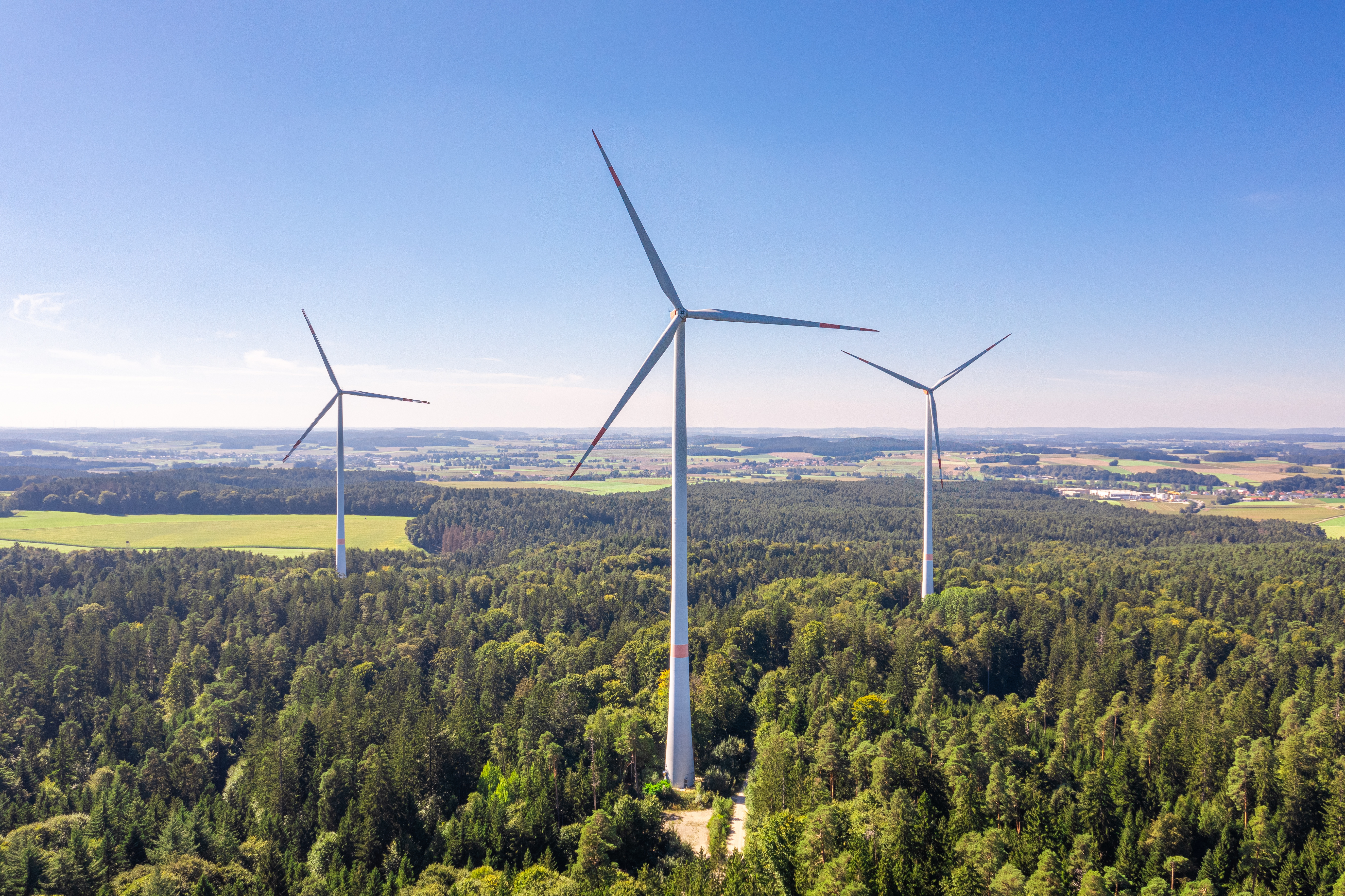 Drei Windräder aus der Luft fotografiert in einem Wald mit Landschaft im Hintergrund.
