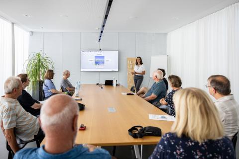 An einem großen Tisch sitzen Personen und hören sich den Vortrag der Stadtwerke Fürth infra zum Thema Photovoltaik an. Im Hintergrund eine Dame und ein Monitor mit den präsentierten Inhalten (Quelle: Daggi Binder, maizucker.de)