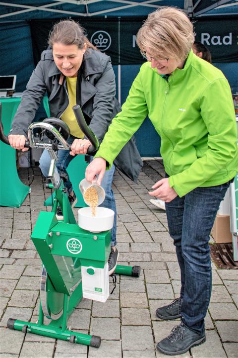 Eine Frau auf einem grünen Ergometer mahlt Getreide mit der eigenen Körperkraft; eine Kollegin schüttet Getreide nach;  (Quelle: Oliver Kordes) 