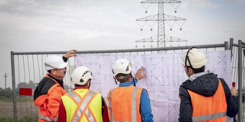 Personen in Warnwesten betrachten einen technischen Plan, im Hintergrund ist ein Strommast (Bildquelle: StMWi).