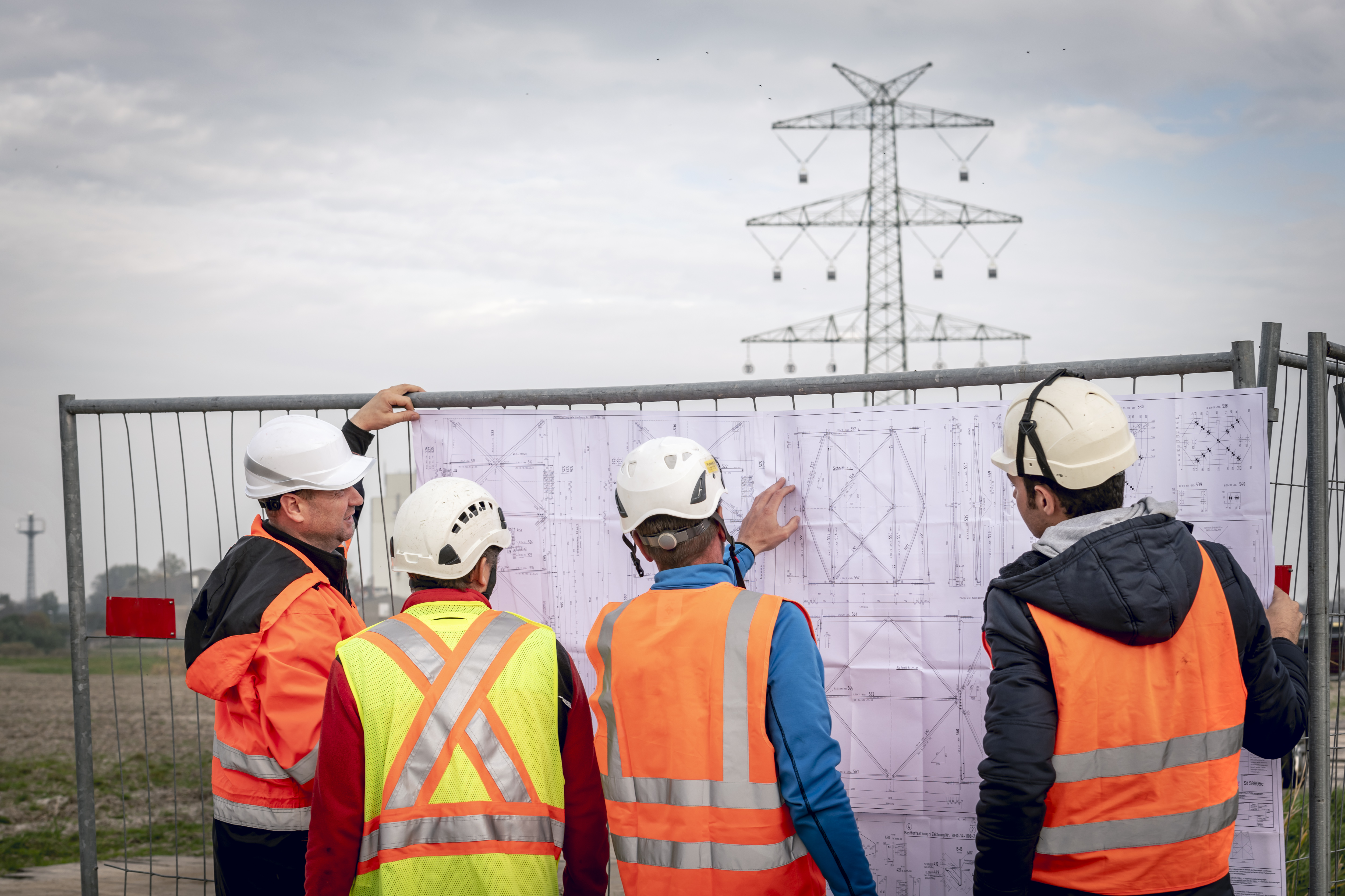 Personen in Warnwesten betrachten einen technischen Plan, im Hintergrund ist ein Strommast (Bildquelle: StMWi).